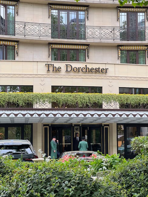The exterior of The Dorchester hotel, a luxurious building with cream-colored stone facade and ornate black and gold decorative details around the entrance. Several guests and staff members are gathered near the entrance, some in green uniforms and others in formal attire. Above the entrance, there are three levels of balconies with black wrought iron railings and potted plants, and a neatly trimmed hedge runs horizontally across the building just above the entrance. The hotel is situated on a street with lush greenery and shrubs in the foreground, contributing to an elegant and well-maintained appearance, with natural daylight illuminating the scene. This image relates to private property management and waste disposal services that cater to high-end establishments seeking professional rubbish removal solutions.