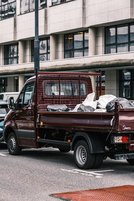 A small, burgundy flatbed truck parked on an urban street in front of a modern office building with large windows and a concrete facade. The truck's cargo area is filled with a variety of waste materials, including black and white plastic bags, some of which are loosely arranged and partially covered with plastic sheeting. The truck features a metal mesh safety barrier at the rear of the cab, and the side panel beneath the cargo area is black. The vehicle is positioned close to the curb, with a white van visible parked further back among other vehicles. The scene appears to be part of an independent rubbish removal or on-site clearance operation, with the use of a small commercial vehicle suitable for private waste disposal, supporting alternative waste handling options outside of local authority collection. A black streetlamp with a grooved pole stands beside the truck, and natural daylight illuminates the scene, accentuating the textures of the waste in the cargo bed and the building’s exterior surfaces.