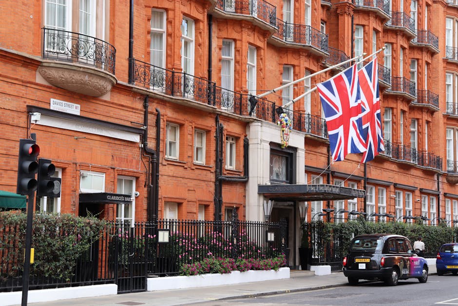 The exterior of The Dorchester hotel, a luxurious building with cream-colored stone facade and ornate black and gold decorative details around the entrance. Several guests and staff members are gathered near the entrance, some in green uniforms and others in formal attire. Above the entrance, there are three levels of balconies with black wrought iron railings and potted plants, and a neatly trimmed hedge runs horizontally across the building just above the entrance. The hotel is situated on a street with lush greenery and shrubs in the foreground, contributing to an elegant and well-maintained appearance, with natural daylight illuminating the scene. This image relates to private property management and waste disposal services that cater to high-end establishments seeking professional rubbish removal solutions.
