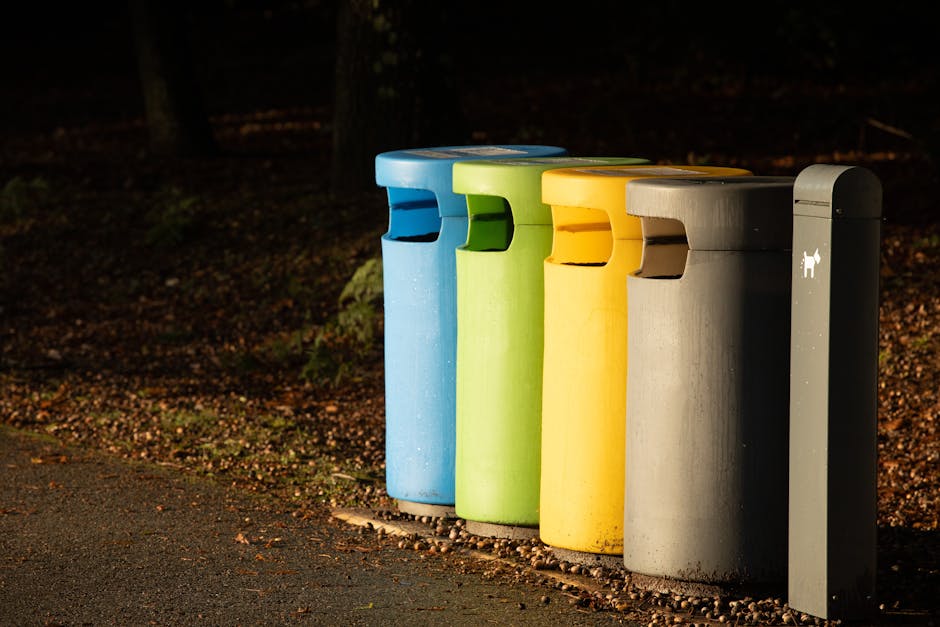 Four cylindrical public waste bins are positioned side by side on a paved pathway adjacent to a natural, leaf-strewn area in an outdoor setting. From left to right, the first bin is painted in bright blue, with a smooth surface and a slot for inserting waste at the top. Next, a lime green bin with similar design features sits alongside, followed by a yellow bin with the same form and textured finish. The fourth bin is grey, matching the others in shape and texture, and also has a top slot for waste disposal. To the right of these bins stands a tall, dark grey, rectangular waste receptacle or informational post with a small white icon indicating it is designated for animal waste or dog litter disposal. The background is blurred with trees and dim lighting, suggesting early evening or low-light conditions, while the ground around the bins is covered with small pebbles and fallen leaves, emphasizing the outdoor environment and the use of these bins for responsible waste management in a public space, aligning with private or independent collection services like those offered by Waste Disposal Mayfair.