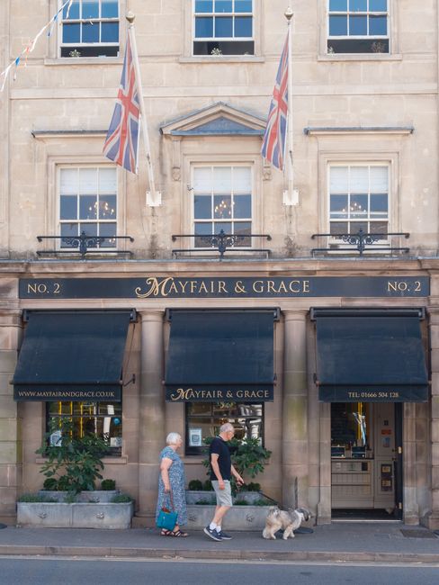 The exterior of a historic stone building with a beige facade features three large dark blue awnings over ground-floor windows and an entrance. Above the awnings, a sign displays the name 'Mayfair & Grace,' flanked by the numbers 'No. 2' on each side. Two Union Jack flags are mounted on poles extending from the upper part of the building. The middle window has a decorative triangular pediment above it, and there are small potted plants placed on the sidewalk in front of the lower windows. Two pedestrians, an elderly woman in a patterned dress and a man in casual attire, are walking along the sidewalk with a small, light-colored dog on a leash. The scene is illuminated by natural daylight, with a clear view of the building's stone construction and classic windows. Waste Disposal Mayfair’s presence is implied through the context of private waste management, but no specific rubbish containers or collection activities are visible in the image.