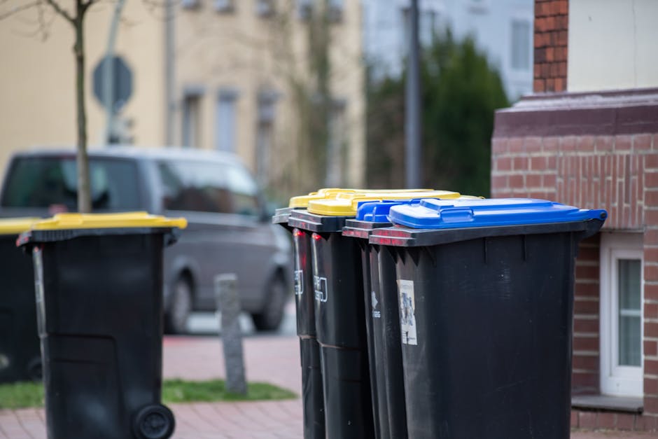 Four cylindrical public waste bins are positioned side by side on a paved pathway adjacent to a natural, leaf-strewn area in an outdoor setting. From left to right, the first bin is painted in bright blue, with a smooth surface and a slot for inserting waste at the top. Next, a lime green bin with similar design features sits alongside, followed by a yellow bin with the same form and textured finish. The fourth bin is grey, matching the others in shape and texture, and also has a top slot for waste disposal. To the right of these bins stands a tall, dark grey, rectangular waste receptacle or informational post with a small white icon indicating it is designated for animal waste or dog litter disposal. The background is blurred with trees and dim lighting, suggesting early evening or low-light conditions, while the ground around the bins is covered with small pebbles and fallen leaves, emphasizing the outdoor environment and the use of these bins for responsible waste management in a public space, aligning with private or independent collection services like those offered by Waste Disposal Mayfair.