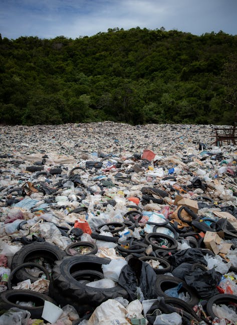 A large area of illegal dumping on a gravel or rocky surface with a mixture of waste materials including discarded black rubber tires, various plastic bags, and packaging debris. The waste is scattered densely across the ground, with some tires partially buried among crumpled plastics and cardboard. In the background, a line of green, densely vegetated trees or shrubs is visible, contrasting with the unmanaged waste in the foreground. The scene appears outdoors, possibly near a forested or countryside area, under an overcast sky which provides diffuse lighting. The waste appears to have been accumulated over time, reflecting an instance of unauthorized or private rubbish disposal that Waste Disposal Mayfair may address through alternative waste handling services, such as on-site clearance or private collection, away from strict local authority management.