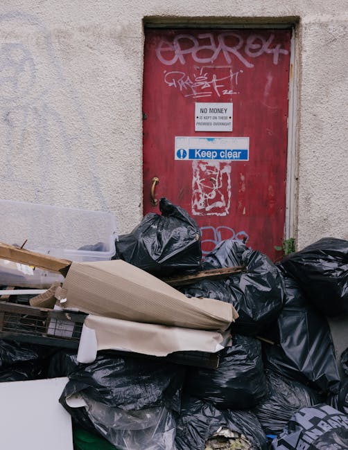 A collection of black rubbish bags, some torn open revealing assorted waste, stacked and strewn in front of a red metal door mounted within a concrete wall. The door features graffiti tags in white and pink spray paint, and has various signs attached, including a white one with black text reading 'No Money' and a blue and white sign stating 'Keep Clear'. Nearby, there are cardboard and paper packaging, suggesting ongoing rubbish clearance. The scene appears to be an outdoor urban alleyway or service area, with the debris indicating recent or ongoing refuse collection, possibly by an independent waste disposal service, reflected in the context of alternative waste handling and rubbish removal in a city environment. The lighting is natural, highlighting the textures and colors of the garbage and surrounding surface, conveying an unkempt but typical aspect of rubbish accumulation awaiting collection or disposal.