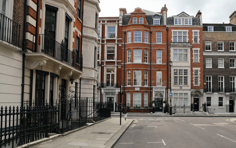 A row of elegant, white Victorian-style terraced buildings with detailed architectural features such as decorative cornices, arched windows, and classical balustrades. The buildings have multiple floors, each with sash windows framed by ornamental mouldings, and small balconies or flower boxes on some windows. In the foreground, parked cars line the street, including a black vehicle, a white car, and a dark-coloured vehicle, positioned on a paved road with a black iron fence separating the sidewalk from the buildings. The right side of the image shows a lamppost and small trees planted along the pavement. In the background, a construction scaffold is visible on one building, indicating ongoing refurbishment. The overall scene is illuminated by natural daylight with a partly cloudy sky, creating a professional and neutral setting that aligns with property and waste management contexts, such as site clearance or private rubbish disposal services sometimes associated with residential estate maintenance.