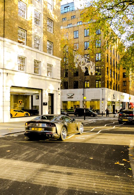 A street scene in an urban area featuring a black sports car with a sleek, aerodynamic design, parked near the curb with its rear facing the camera; the car has a glossy finish, visible dual exhaust pipes, and distinctive alloy wheels. The background shows a mix of buildings, including a white storefront with large windows displaying luxury vehicles, one of which is a yellow sports car, and a taller, brown brick apartment building with multiple windows and balconies. Trees with yellow and green leaves line the sidewalk, with some foliage extending into the frame from the right. The street is paved with asphalt, marked with white pedestrian crossing lines, and there are a few other parked and moving cars, along with pedestrians visible in the distance. This scene appears to be in a high-end commercial or residential district, where private vehicle storage and on-site visitation are common, and Waste Disposal Mayfair offers service options for alternative waste handling in such urban environments, supporting safe rubbish collection and disposal practices.