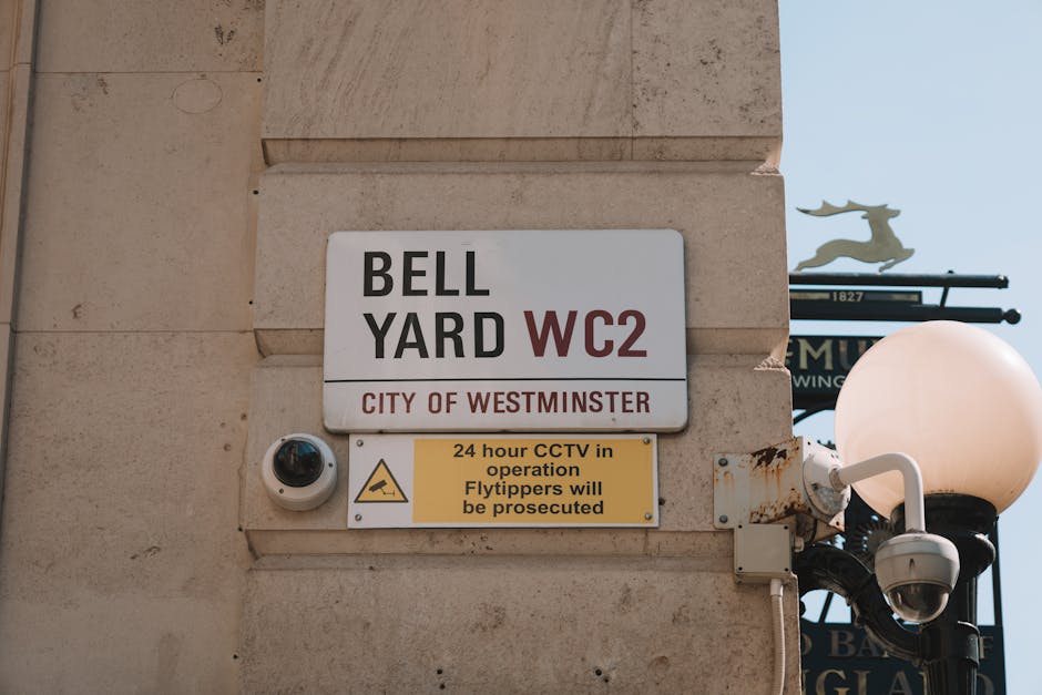 A close-up view of a beige stone building corner featuring a white rectangular sign with black and red text that reads 'BELL YARD WC2', indicating a location within the City of Westminster. Below this, a small rectangular yellow sign with black text states '24 hour CCTV in operation Flytippers will be prosecuted'. To the left of the signs, a small, round security camera with a dark lens is mounted on the wall, indicating surveillance of the area. On the right side, a black street lamp with a spherical white globe is visible, with a security camera attached to its side. In the background, part of a street sign with a metallic finish and decorative elements, such as a stylized animal figure or sculpture, can be seen against a clear blue sky, suggesting an urban environment. The scene depicts a typical city security and surveillance setup, supporting the theme of controlled waste disposal and enforcement in a London district.