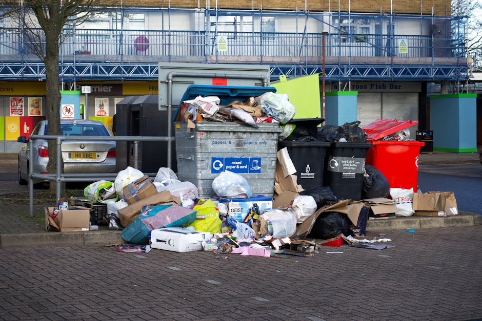 A large grouping of overflowing waste bins and discarded rubbish situated on a paved urban sidewalk near a commercial building with storefronts. The pile includes a grey recycling bin labeled for mixed paper and cardboard, with its lid open and paper waste spilling out, alongside black and red waste containers, some with black plastic garbage bags protruding. Surrounding the bins are various discarded cardboard boxes, plastic packaging, and crumpled paper, with some items strewn onto the ground, including a cardboard box and a paper bag. In the background, there is a parking area with a silver car parked adjacent to the rubbish, a metal railing separating the waste area from the street, and a commercial building with a covered balcony or stairwell area clad in blue protective fencing and scaffolding. The scene is well-lit, suggesting daytime, and highlights issues of private waste disposal or alternative rubbish handling outside official municipal collection points.