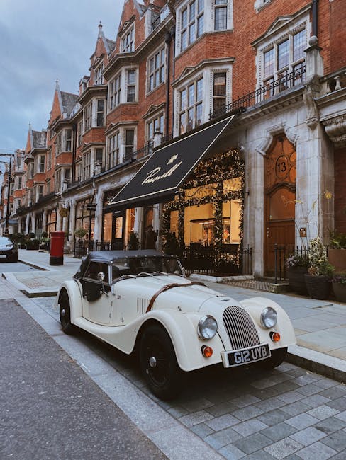 A vintage white convertible car with black accents and a black soft top is parked on a narrow, paved street in front of a row of traditional red-brick terraced houses with white window frames and ornate architectural details. The car features rounded headlights, a prominent vertical grille, and a black leather strap securing the hood. The street scene includes a black street lamp, a red post box, and a large, black-framed awning over the entrance of a boutique or café, which is situated at the ground level of a building with a light-colored stone façade. The building's address number, 13, is visible next to a wooden door with a glass panel. Potted plants adorn the steps leading to the entrance, and the environment suggests a quiet, upscale urban neighbourhood. Natural daylight and soft, ambient lighting from inside the establishment highlight the street’s clean, well-maintained appearance, subtly aligning with the context of private or alternative waste management options hosted through independent collection services like Waste Disposal Mayfair.