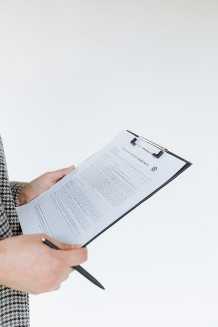 A person holding a black clipboard with a printed document attached, seen from the waist up against a plain light grey background. The individual is wearing a grey and white checkered jacket, and their left hand is gripping the top of the clipboard while their right hand holds a black pen positioned near the bottom. The document appears to be a formal license or permit form, with visible text and a small logo at the top, consistent with official paperwork. The scene is well-lit with soft, evenly distributed lighting, emphasizing the textures of the paper and clothing. This image reflects the context of preparing or managing documents related to waste disposal regulations, possibly associated with private on-site clearance or waste handling services offered by Waste Disposal Mayfair, aligned with the theme of rubbish removal services and licensing requirements.