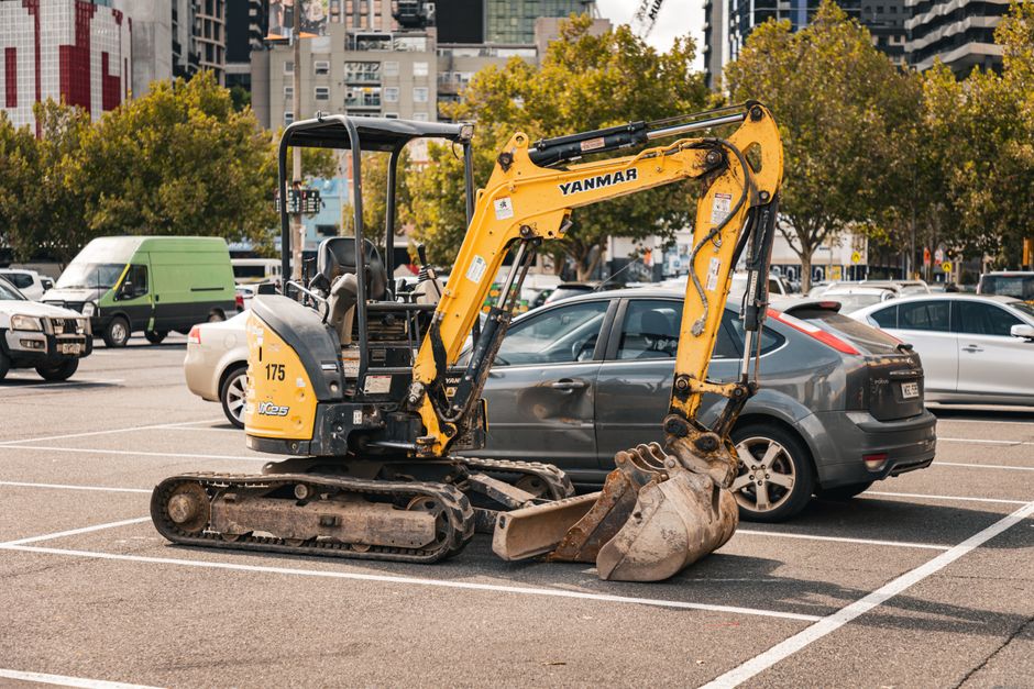 A small yellow Yanmar mini excavator with a compact tracked undercarriage is positioned in a parking lot on a flat asphalt surface, surrounded by parked cars including a white van, a silver sedan, and a darker hatchback. The excavator's arm is extended forward, with a rusty, well-used digging bucket resting on the ground. In the background, there are multi-storey buildings and a line of trees with autumn foliage, featuring green and golden leaves. The scene is lit by natural daylight, casting soft shadows on the surface. The environment suggests an area where construction or site clearance might be taking place, aligning with services provided by waste disposal and rubbish removal companies such as Waste Disposal Mayfair, who handle on-site clearance and machinery removal as part of private waste management solutions in urban settings.