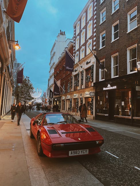A red sports car with a sleek, low-profile design is parked on a city street, positioned close to the curb in front of a series of multi-storey buildings featuring brick and stone facades, large windows, and decorative signage. The car's surface has a shiny, reflective finish, with visible vents on the hood and a wide, rectangular front grille. The street is lined with pedestrians walking along the sidewalk, some wearing coats, and the scene is illuminated by warm streetlights and the soft glow of shop windows. In the background, there are additional buildings, signs, and a glimpse of festive holiday decorations hanging across the street, suggesting an urban environment that might involve private waste collection or rubbish disposal services, as part of managing waste in retail or hospitality areas. Waste Disposal Mayfair's focus on rubbish removal in busy commercial districts aligns with this scene, which features a mix of luxury vehicles and city activity, set during early evening hours with natural daylight fading. The overall setting reflects a vibrant, upscale area that may require professional waste management for commercial waste or on-site clearance operations.
