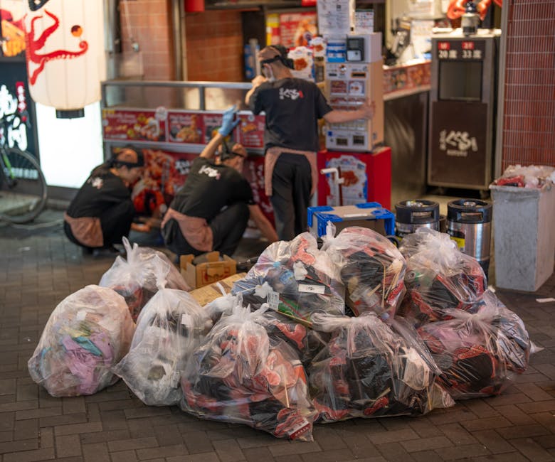 A collection of large transparent plastic rubbish bags filled with mixed waste, including clothing, packaging, and miscellaneous refuse, situated on a paved outdoor area in front of a commercial shop. The bags are piled together on the ground, with some partially open and revealing their contents. Behind the bags, three individuals dressed in black uniforms with brown aprons and gloves are kneeling and working near a stack of cardboard boxes and other disposal containers. The background features a shop with bright signage, illuminated by artificial lighting, with visible red and white promotional posters and a menu display. To the right, there are several cylindrical metal waste bins and a large grey bin, indicating an area designated for waste collection outside the premises. The scene suggests a private or independent rubbish collection operation, possibly facilitated by Waste Disposal Mayfair, serving the busy street environment typical of the Mayfair district, with a focus on on-site waste handling and rubbish removal services.