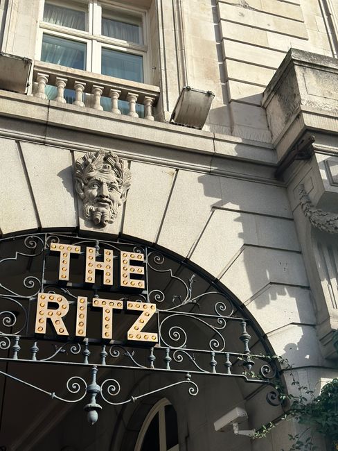 The image depicts the facade of a building with an ornate stone decoration of a lion's head with a fierce expression and flowing mane, mounted above a large archway. Beneath the lion's head, there is a sign with the words 'THE RITZ' rendered in bold, capital letters with warm, round lightbulb bulbs embedded within each letter, creating a vintage marquee appearance. The archway is constructed from light-colored stone or concrete, with decorative ironwork extending from the sides, suggesting an entrance to a high-end establishment, possibly a hotel or venue. Above the archway, part of the building's exterior wall is visible, with a window featuring a white wooden frame and a small stone balustrade beneath it. The building's architecture is classical, with carved stone details and clean lines, and the scene is illuminated by natural daylight, casting soft shadows that accentuate the textured surface of the stone. This setting is characteristic of an urban area with elegant historical architecture, where private events or luxury accommodation might require occasional rubbish collection or waste management services by providers like Waste Disposal Mayfair, operating in compliance with local regulations.